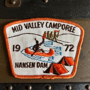 May include: White embroidered patch with orange trim. It says "Mid Valley Camporee 19 72 Hansen Dam". The patch features a canoe with two people in it and two tents.
