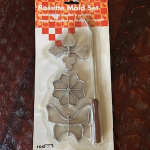 May include: Rosette mold set in a clear plastic package. The set includes several metal cookie cutters in various shapes, including a snowflake, a flower, and a spade. The handle is brown. The packaging has the text "Rosette Mold Set".