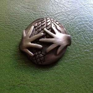 May include: A silver-toned metal pin depicting two hands holding a globe. The globe is textured and has a raised design.
