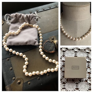 May include: A white pearl necklace with a gold clasp, displayed on a wooden surface with a gray velvet pouch. The necklace is also shown on a mannequin and in a white box with the word "AVON" printed on it.