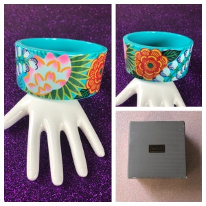 May include: A turquoise bangle bracelet with a floral design in shades of pink, orange, green, and white. The bracelet is in a box with the word "AVON" printed on it.