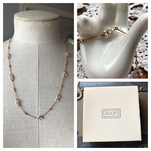 May include: A gold chain necklace with small white pearls. The necklace is displayed on a white mannequin. The necklace comes in a white box with the word "AVON" printed on it.