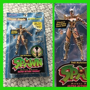 May include: A plastic action figure of Cosmic Angela from the Spawn comic book series. The figure is wearing a silver and gold costume with a black and white animal print design. The figure has wings and is holding a sword. The figure is packaged in a plastic blister pack with a Spawn logo and the words "Deluxe Edition Ultra-Action Figures".