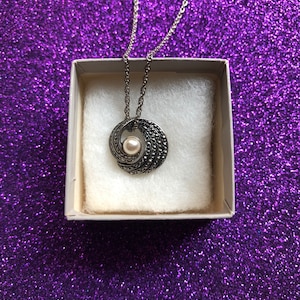 May include: A silver-toned necklace with a circular pendant featuring a central pearl and intricate detailing. The necklace is presented in a small, white box with a soft, white interior, set against a sparkling purple background.