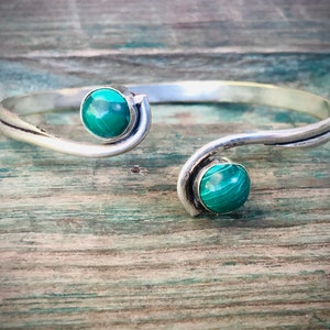 May include: A silver bangle bracelet with two green malachite stones. The bracelet is open and has a curved design.