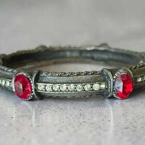Peut inclure: Une bague circulaire avec une finition argentée oxydée comportant deux pierres rouges taille ovale placées à intervalles. L'anneau est orné d'une rangée de petits cristaux clairs incrustés entre les pierres rouges et de détails métalliques texturés.