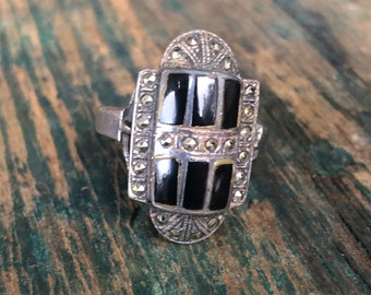 May include: Silver-toned ring with a rectangular black onyx center, flanked by smaller rectangular onyx stones. The ring is accented with small, round, silver-colored stones. The ring has an Art Deco design.