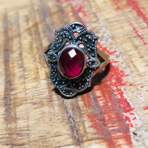 May include: A silver ring with a large, oval, faceted, red gemstone at the center. The ring has an ornate, filigree design with small, round, dark accents. The ring is set against a wooden surface.