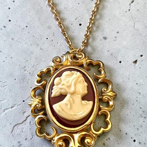 Pode incluir: Um colar com pingente de camafeu dourado. O camafeu oval apresenta um perfil creme sobre um fundo vermelho escuro, emoldurado por uma moldura dourada decorativa. O colar tem uma delicada corrente dourada.