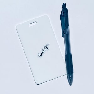 May include: A white rectangular tag with a slot at the top and the words "Thank you" in cursive. A black and clear pen is next to the tag. The pen has a black grip and a clear barrel.