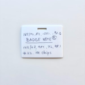 Puede incluir: Una placa rectangular de pl&aacute;stico blanco con una ranura horizontal en la parte superior. La superficie presenta texto escrito a mano con tinta azul: 165/72, 82, 100%, 97.6, BADGE NOTE, 144/67, 98%, 72, 98.1, #22. ice chips. La placa tiene un acabado mate liso.