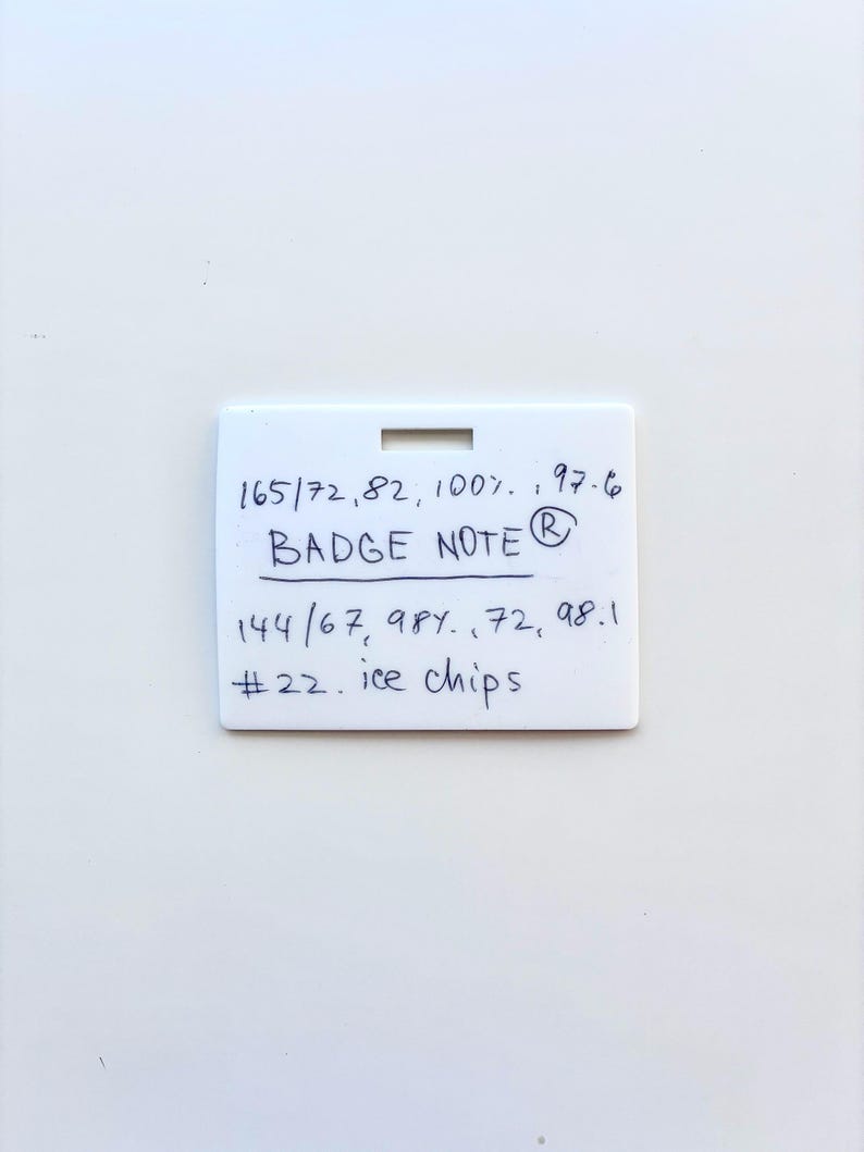 Puede incluir: Una placa rectangular de pl&aacute;stico blanco con una ranura horizontal en la parte superior. La superficie presenta texto escrito a mano con tinta azul: 165/72, 82, 100%, 97.6, BADGE NOTE, 144/67, 98%, 72, 98.1, #22. ice chips. La placa tiene un acabado mate liso.