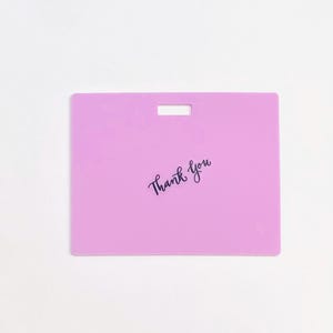 Puede incluir: Una etiqueta rectangular de pl&aacute;stico de color morado claro con una ranura horizontal en la parte superior central para su fijaci&oacute;n. Las palabras Thank You est&aacute;n impresas en una fuente cursiva negra en el centro de la superficie plana.