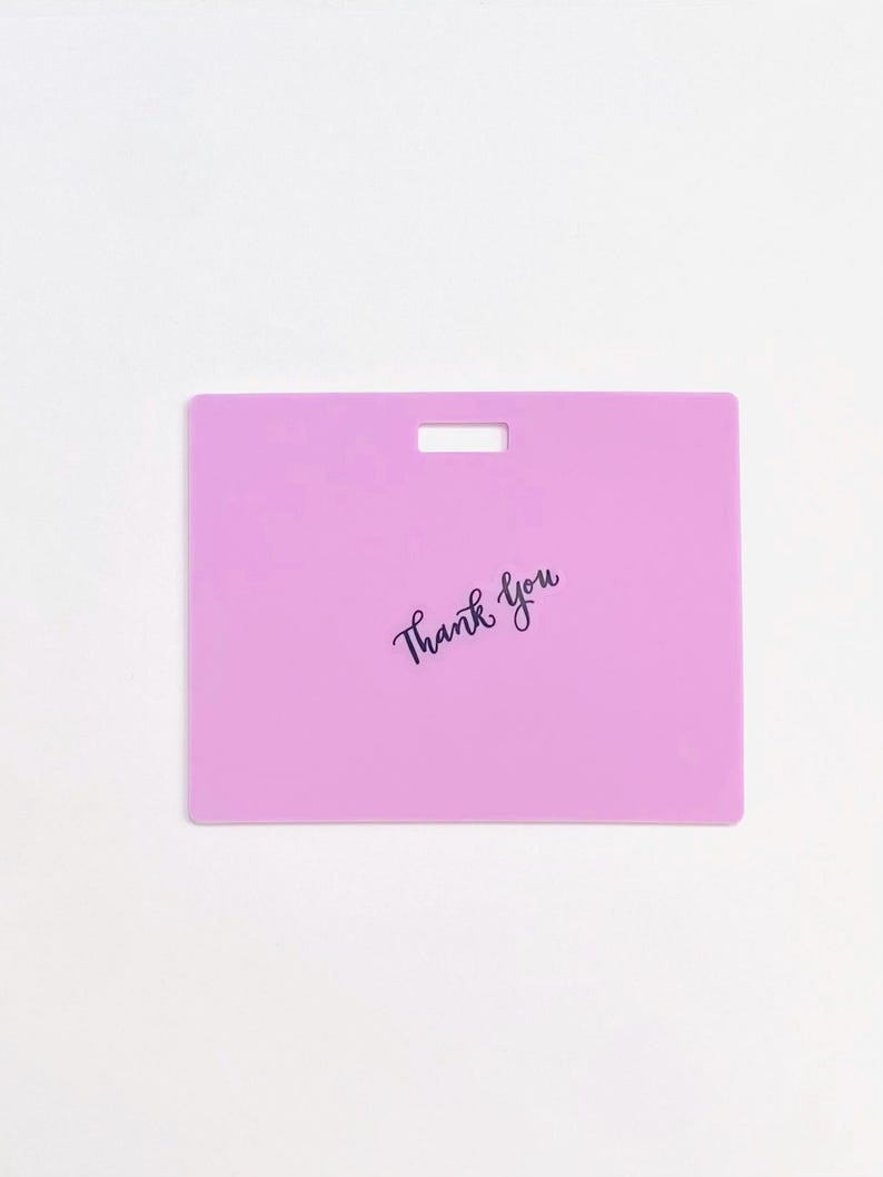 Puede incluir: Una etiqueta rectangular de pl&aacute;stico de color morado claro con una ranura horizontal en la parte superior central para su fijaci&oacute;n. Las palabras Thank You est&aacute;n impresas en una fuente cursiva negra en el centro de la superficie plana.