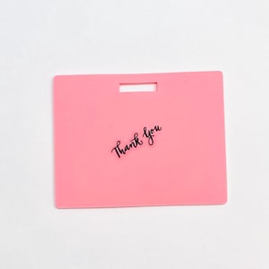 Puede incluir: Una etiqueta rectangular de pl&aacute;stico rosa con una ranura horizontal en la parte superior central para su fijaci&oacute;n. Las palabras Thank You est&aacute;n impresas en una fuente cursiva negra en el centro de la etiqueta, que tiene una superficie lisa.