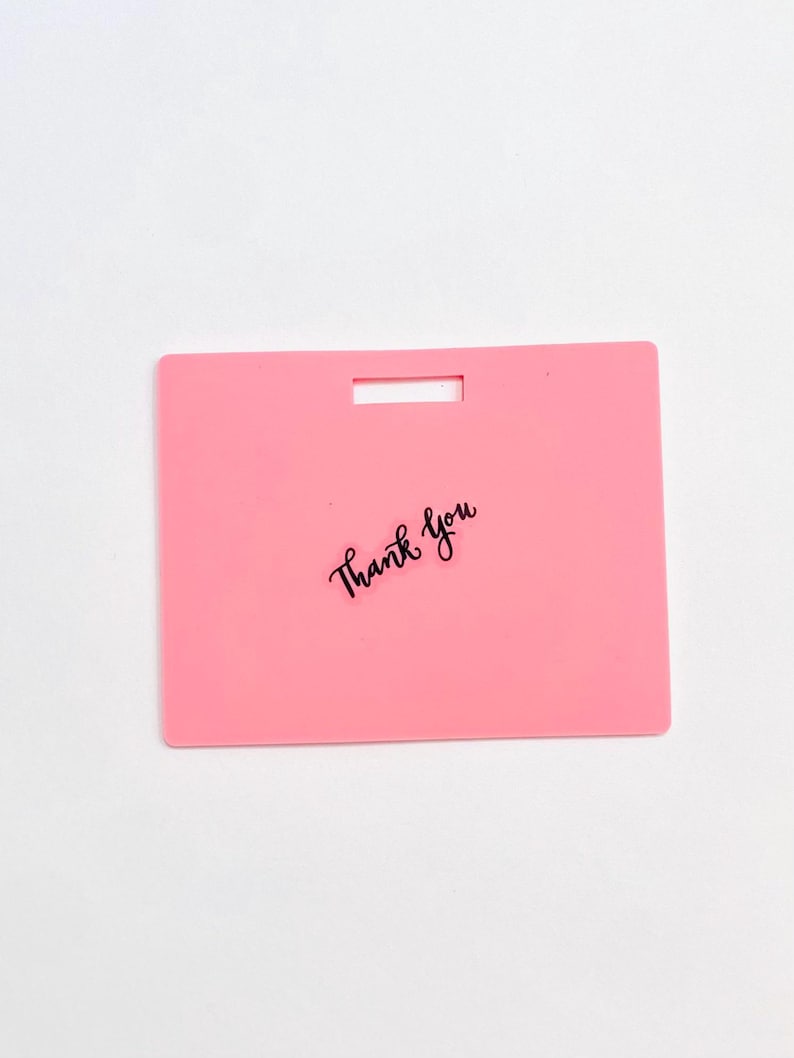 Puede incluir: Una etiqueta rectangular de pl&aacute;stico rosa con una ranura horizontal en la parte superior central para su fijaci&oacute;n. Las palabras Thank You est&aacute;n impresas en una fuente cursiva negra en el centro de la etiqueta, que tiene una superficie lisa.