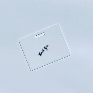 Puede incluir: Una etiqueta rectangular de acr&iacute;lico blanco con una ranura horizontal en la parte superior central y las palabras Thank you impresas en una fuente cursiva negra, colocadas en &aacute;ngulo en la parte inferior izquierda.