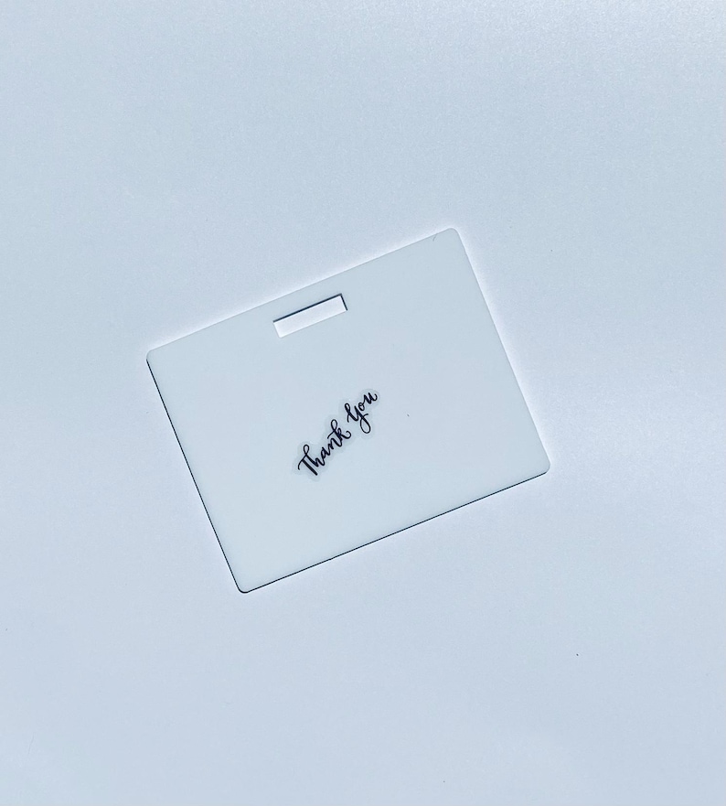 Puede incluir: Una etiqueta rectangular de acr&iacute;lico blanco con una ranura horizontal en la parte superior central y las palabras Thank you impresas en una fuente cursiva negra, colocadas en &aacute;ngulo en la parte inferior izquierda.