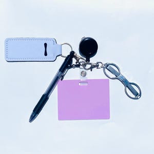 Könnte beinhalten: Eine Sammlung von Accessoires, darunter ein helllila Ausweis, eine schwarze, einziehbare Ausweisrolle, ein Stift und ein silberner Brillenhalter. Die Artikel sind an einem Schlüsselring befestigt und vor weißem Hintergrund.