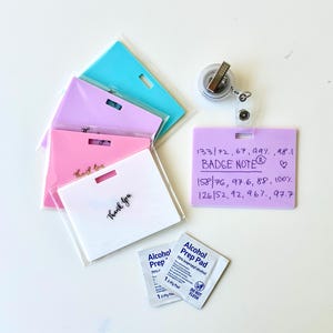 Può includere: Un set di porta badge rettangolari in plastica nei colori bianco, rosa, lavanda e azzurro, con un porta badge color lavanda che riporta segni vitali medici scritti a mano e il testo BADGE NOTE. Include un avvolgitore per badge e due salviette.
