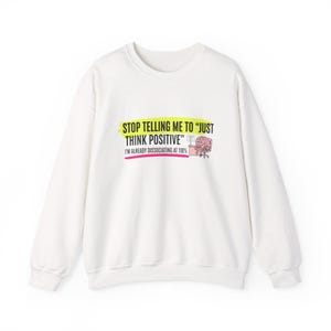 May include: White sweatshirt with the text "STOP TELLING ME TO 'JUST THINK POSITIVE'" in yellow and black. Below the text is "I'M ALREADY DISSOCIATING AT 110%" in red. A graphic of a brain at a desk is also included.
