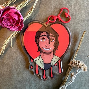May include: A heart-shaped acrylic keychain featuring a cartoon illustration of a person with brown hair and a light green shirt. The background of the illustration is a solid red heart with red drips at the bottom. It includes a red heart-shaped clasp.