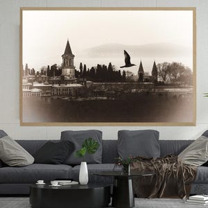 May include: Sepia-toned framed print of a historic cityscape with a flying bird. The artwork hangs above a dark gray sofa with decorative pillows. A modern lamp and potted plant complete the living room setting.