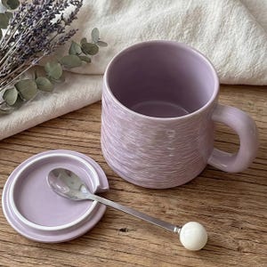 Puede incluir: Una taza de cerámica lila con tapa a juego y una cuchara pequeña. La taza tiene una superficie texturizada y un asa redondeada. La cuchara tiene una bola blanca en el extremo del mango. Lavanda seca y un paño blanco en el fondo.