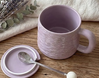 May include: A lavender ceramic mug with a matching lid and a small spoon. The mug has a textured surface and a rounded handle. The spoon has a white ball at the end of the handle. Dried lavender and a white cloth are in the background.
