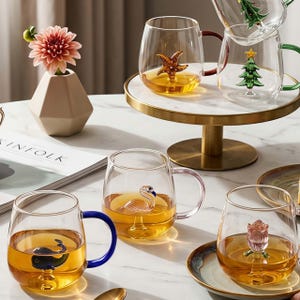 May include: Glass mugs with coloured handles and decorative sea creatures and a Christmas tree inside. The mugs are filled with a golden liquid, and one mug has a pink flamingo. A small vase with a pink flower is also present.
