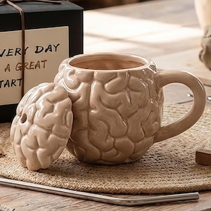Puede incluir: Una taza de cerámica beige con forma de cerebro humano, con una tapa a juego. La taza tiene un asa y está sobre una esterilla tejida. El texto en la caja dice "EVERY DAY A GREAT START".
