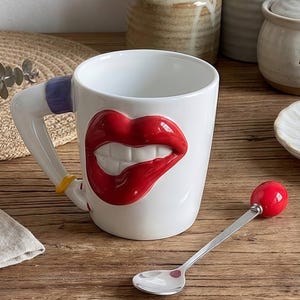 Puede incluir: Taza de cerámica blanca con un diseño 3D de labios rojos y dientes blancos. El asa tiene detalles azules y amarillos. Una cuchara plateada con una bola roja descansa junto a la taza. Sobre una superficie de madera.