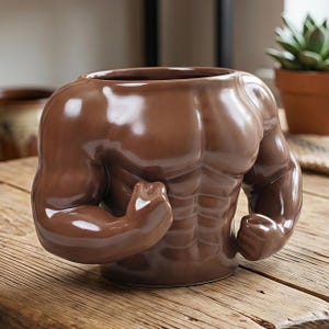 Puede incluir: Una taza de cerámica marrón con forma de torso musculoso con brazos flexionados. La taza tiene un acabado brillante y está diseñada para contener bebidas o servir como objeto decorativo. Está sobre una superficie de madera.