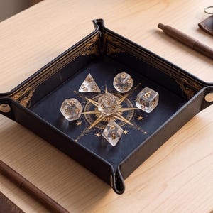 May include: A square black folding dice tray with gold-coloured metal snaps at the corners. The interior features a gold compass rose design printed on a dark background, containing five clear polyhedral dice with gold-coloured numbers.