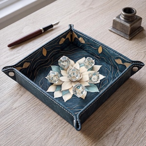 Puede incluir: Una bandeja cuadrada para dados de cuero azul oscuro con esquinas de botones a presión, con un diseño impreso de una flor de loto blanca y azul claro sobre un fondo azul oscuro con detalles en pan de oro, conteniendo cinco dados poliédricos.