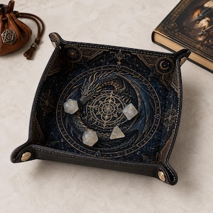 May include: A square, black leather dice tray with gold-colored metal snaps at the corners. The interior features a detailed print of a coiled dragon surrounding a central geometric compass design in shades of blue and gold. Four translucent dice rest inside.
