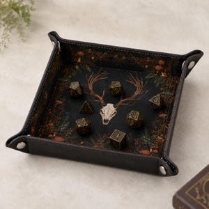May include: A square, dark brown leatherette dice tray with silver-tone metal snaps at the corners. The interior features a printed design of a deer skull centred among dark forest foliage, mushrooms, and ferns, holding six metallic polyhedral dice.