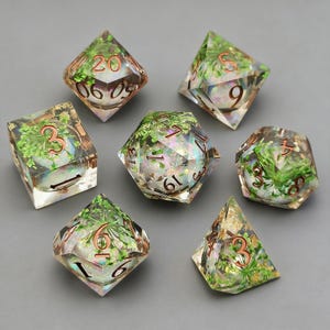 May include: A set of seven polyhedral dice for tabletop games. Each die is made of clear resin, with green plant inclusions and copper-coloured numbering. The dice are various shapes, including a d20, d12, d10, d8, d6, and d4. Ideal for role-playing games.