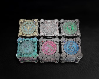 May include: Six square, metallic boxes with detailed designs. Each box has a central circular emblem with symbols and colours, including teal, green, and blue. The boxes are arranged in a grid pattern against a dark background.