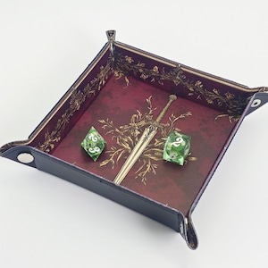 May include: A square, foldable dice tray with a dark maroon interior featuring a gold sword and floral illustration. The tray has black exterior sides secured with silver metal snaps at each corner. Two translucent green dice with white numbers sit inside.
