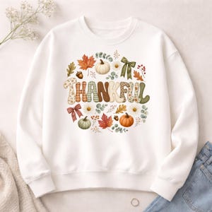 Peut inclure: Un sweat-shirt blanc à col rond orné d'un motif central avec le mot THANKFUL en grandes lettres à motifs. Le texte est entouré d'illustrations de citrouilles, de feuilles d'automne, de glands, de marguerites, de brins d'eucalyptus et de deux nœuds.