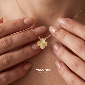 May include: A gold-toned necklace featuring a four-leaf clover pendant with a textured, ribbed surface and a small central bead. The pendant is suspended from a delicate gold-toned chain. The text VELORIQ NEW YORK is printed at the bottom.