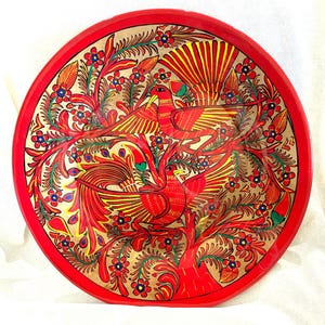 May include: A round, red decorative tray with a hand-painted design. The tray features a gold background with red birds, flowers, and foliage. The birds have outstretched wings, and the flowers are blue and red.