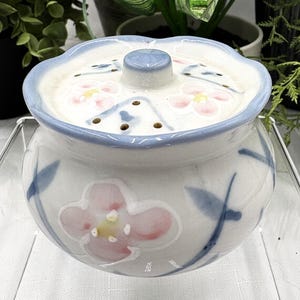 May include: A white ceramic jar with a matching lid featuring a scalloped edge and a central knob. The jar and lid are decorated with hand-painted pink flowers, yellow centers, and blue leaves. The lid includes a series of small circular perforations.