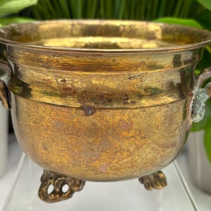 May include: A round brass pot with a weathered, golden-brown patina, featuring two small side handles and three ornate, scrolled feet. The metal surface shows visible signs of oxidation and wear, with a wide, open top rim.