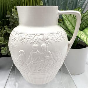 May include: A matte white ceramic pitcher featuring a raised relief design of two figures sitting with a dog in a landscape. The upper portion displays a grape vine motif, while the lower section has vertical fluting. It has a single handle.