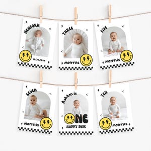 May include: A collection of six baby milestone cards suspended from a string. Each card displays a baby's photo and text denoting the baby's age in months, with a smiley face and lightning bolt design. The cards are secured by wooden clips.