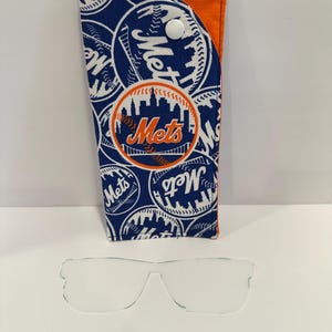 May include: A blue and orange Mets-themed glasses case with a white snap closure. The case features a repeating pattern of the Mets logo and the word "Mets". Clear eyeglasses are in front of the case.