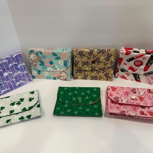 以下が含まれることがあります： さまざまな柄の布製財布のコレクション。デザインには、トンボ、ナマケモノ、クマ、口紅、クローバー、ハート、花柄などがあります。各財布には、カーブしたフラップとボタン留めが付いています。