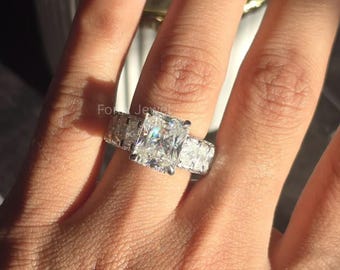 Puede incluir: Un anillo de tono plateado con una gran piedra central rectangular de corte radiante, flanqueada por dos piedras laterales rectangulares más pequeñas en la banda. El engaste de metal es pulido y reflectante, captando la luz para resaltar las piedras claras y facetadas.